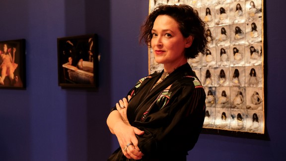 Portrait of curator and writer Hettie Judah, standing with arms crossed in front of an artwork on a purple wall.